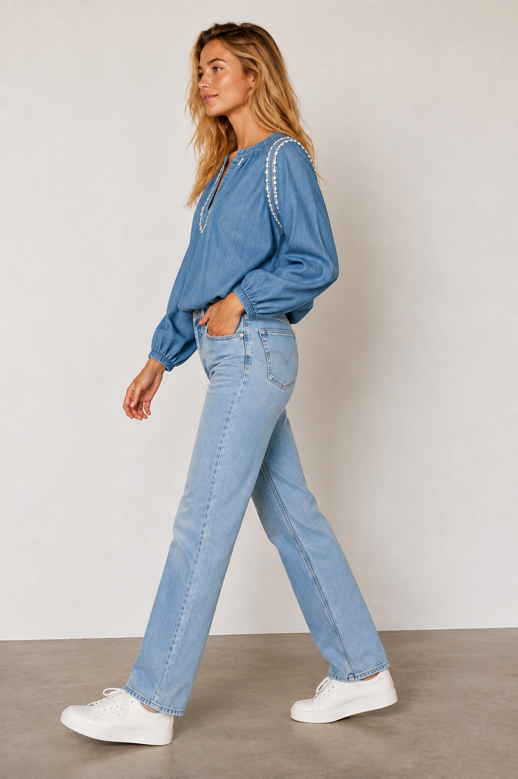 A woman modeling a light blue long-sleeve top with white beaded trim along the neckline and sleeves, paired with light blue jeans and white sneakers, set against a neutral studio background.