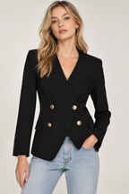 A woman wearing a black double-breasted blazer with gold buttons and light blue jeans, standing against a light gray studio background.