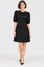 A woman modeling a black short puff-sleeve dress with a gathered neckline and flared skirt, paired with black pointed heels, against a light gray studio background.