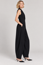 A woman modeling a black sleeveless wide-leg jumpsuit with side pockets, paired with black pointed-toe heels, against a light gray studio background.