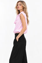 A woman modeling a sleeveless light pink blouse with a bow tie neckline, paired with black wide-leg trousers, against a plain white studio background.