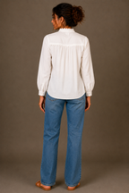 A woman viewed from the back wearing a white long-sleeve blouse with a gathered, ruffled yoke and blue jeans, standing against a solid taupe studio background.