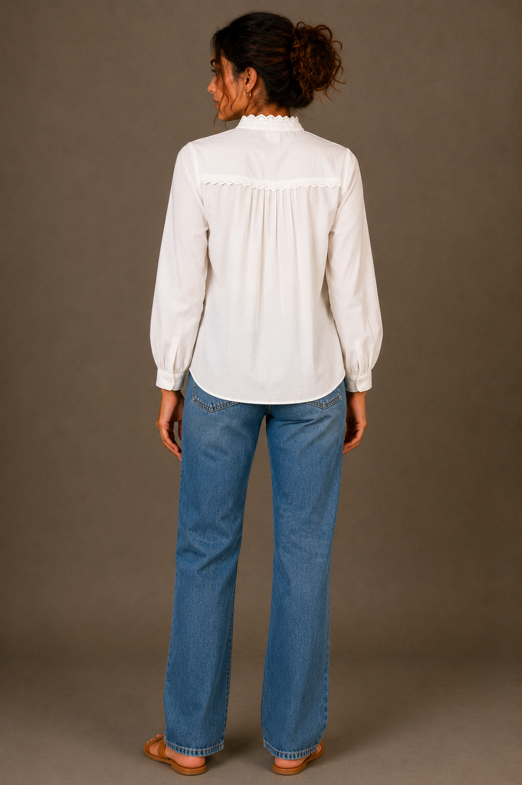 A woman viewed from the back wearing a white long-sleeve blouse with a gathered, ruffled yoke and blue jeans, standing against a solid taupe studio background.