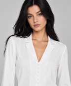 A woman with long dark hair wearing a white long-sleeve button-down blouse with notched lapels, photographed against a neutral light gray studio background.