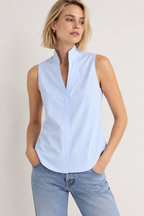 A woman wearing a light blue sleeveless button-front blouse with a stand collar and light-wash blue jeans, photographed against a plain light beige studio background.