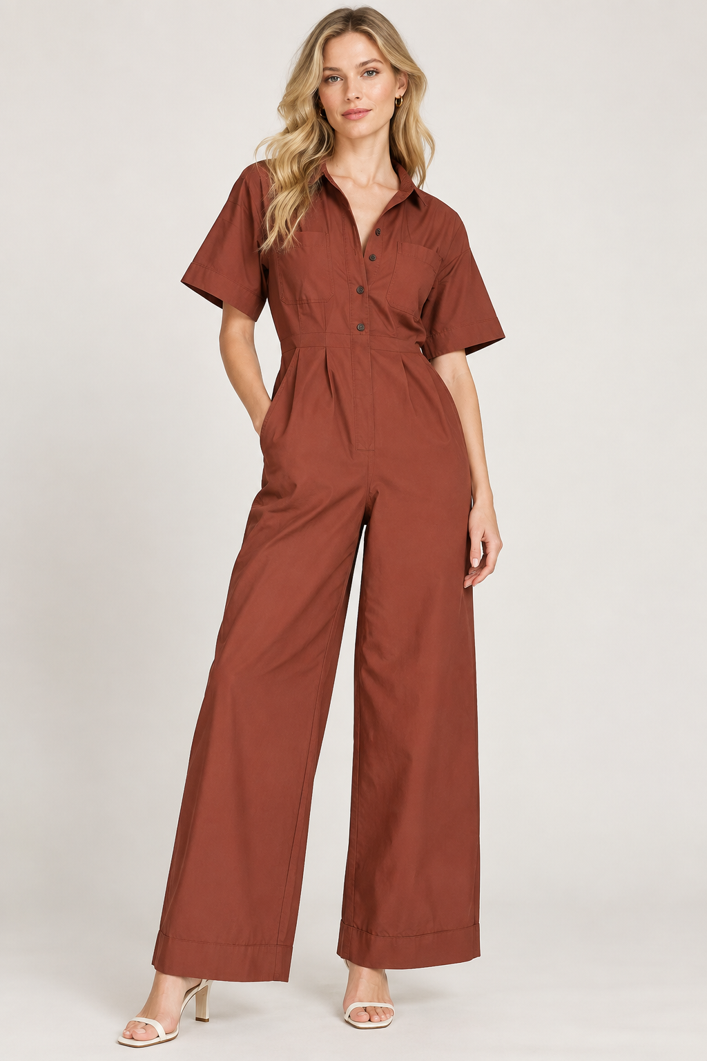 A woman modeling a rust-brown short-sleeve jumpsuit with a button-down front and wide legs, paired with white heeled sandals, against a light gray studio background.