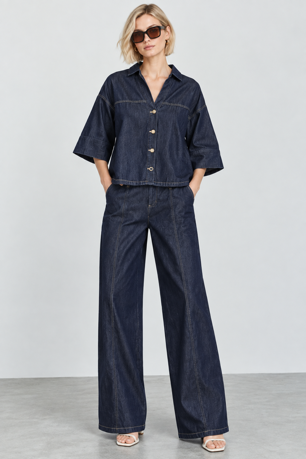 A woman modeling a dark indigo denim two-piece set consisting of a short-sleeve button-front shirt and matching wide-leg pants, styled with sunglasses and flat sandals against a light gray studio background.