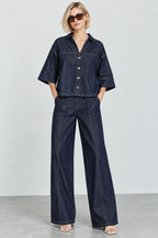 A woman modeling a dark indigo denim two-piece set consisting of a short-sleeve button-front shirt and matching wide-leg pants, styled with sunglasses and flat sandals against a light gray studio background.
