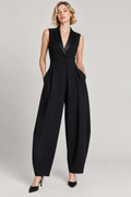 A woman modeling a sleeveless black jumpsuit with a structured collar, single front button, and wide-leg trousers, photographed against a plain light gray studio background.