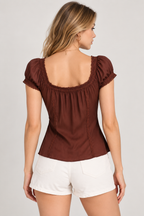 A woman shown from the back wearing a brown short-sleeve top with a square neckline, ruffled trim, and gathered waist, paired with white shorts, against a light gray studio background.