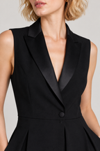 Close-up of a model wearing a black sleeveless blazer-style dress with satin lapels and a single front button, against a light gray studio background.