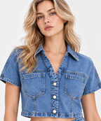 A woman wearing a cropped blue denim shirt jacket with short sleeves, a pointed collar, and front button closure, photographed against a clean light gray studio background.
