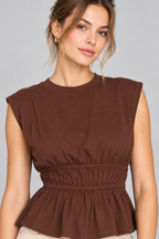 A woman wearing a sleeveless brown peplum top with a gathered elastic waist, shown against a plain light gray studio background.