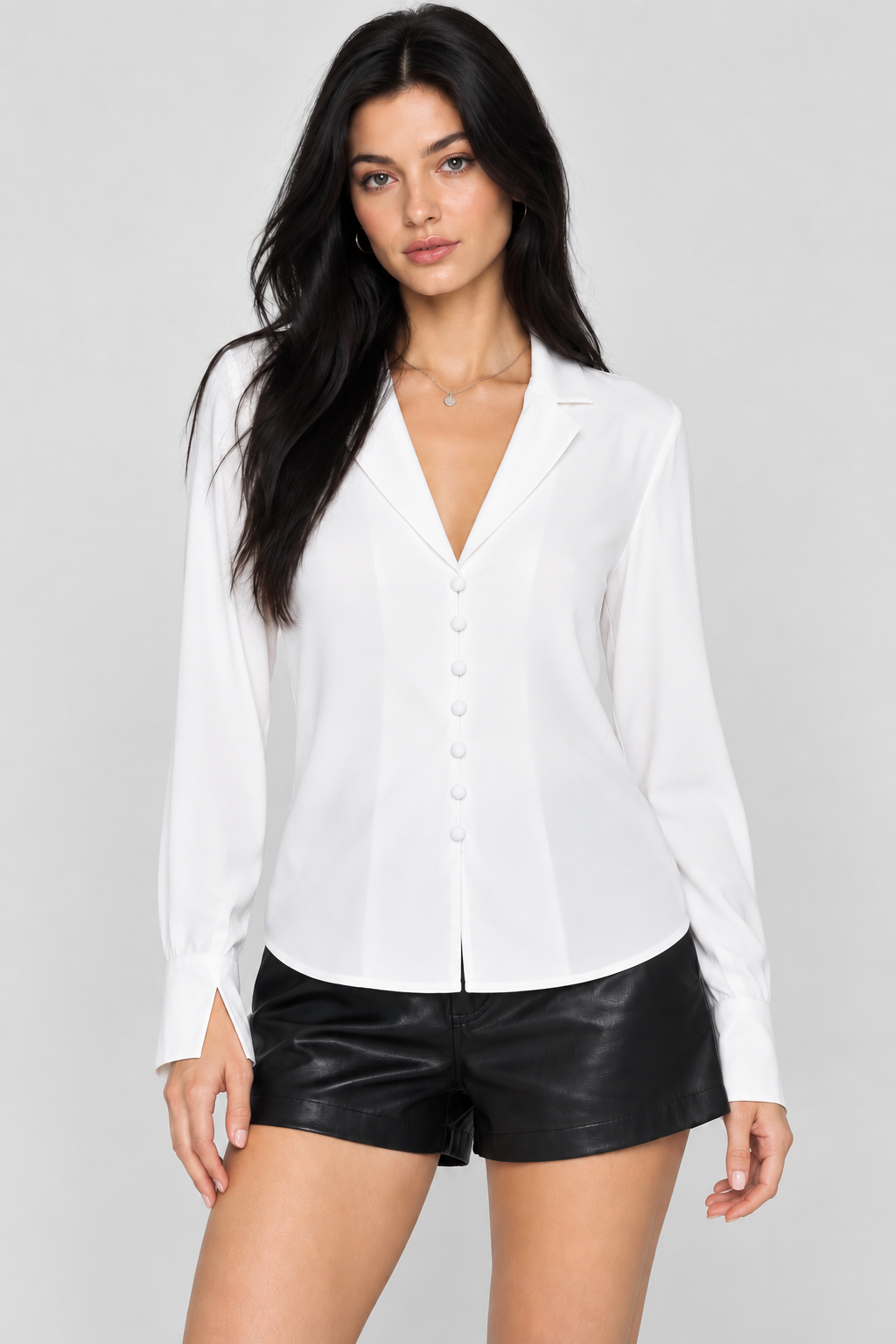 A woman modeling a white long-sleeve button-front blouse with notched lapels, paired with black faux-leather shorts, against a light gray studio background.