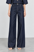 A model wearing dark blue wide-leg denim jeans with a button fly and contrast stitching, paired with a matching dark blue top and white heeled sandals, set against a neutral studio background.