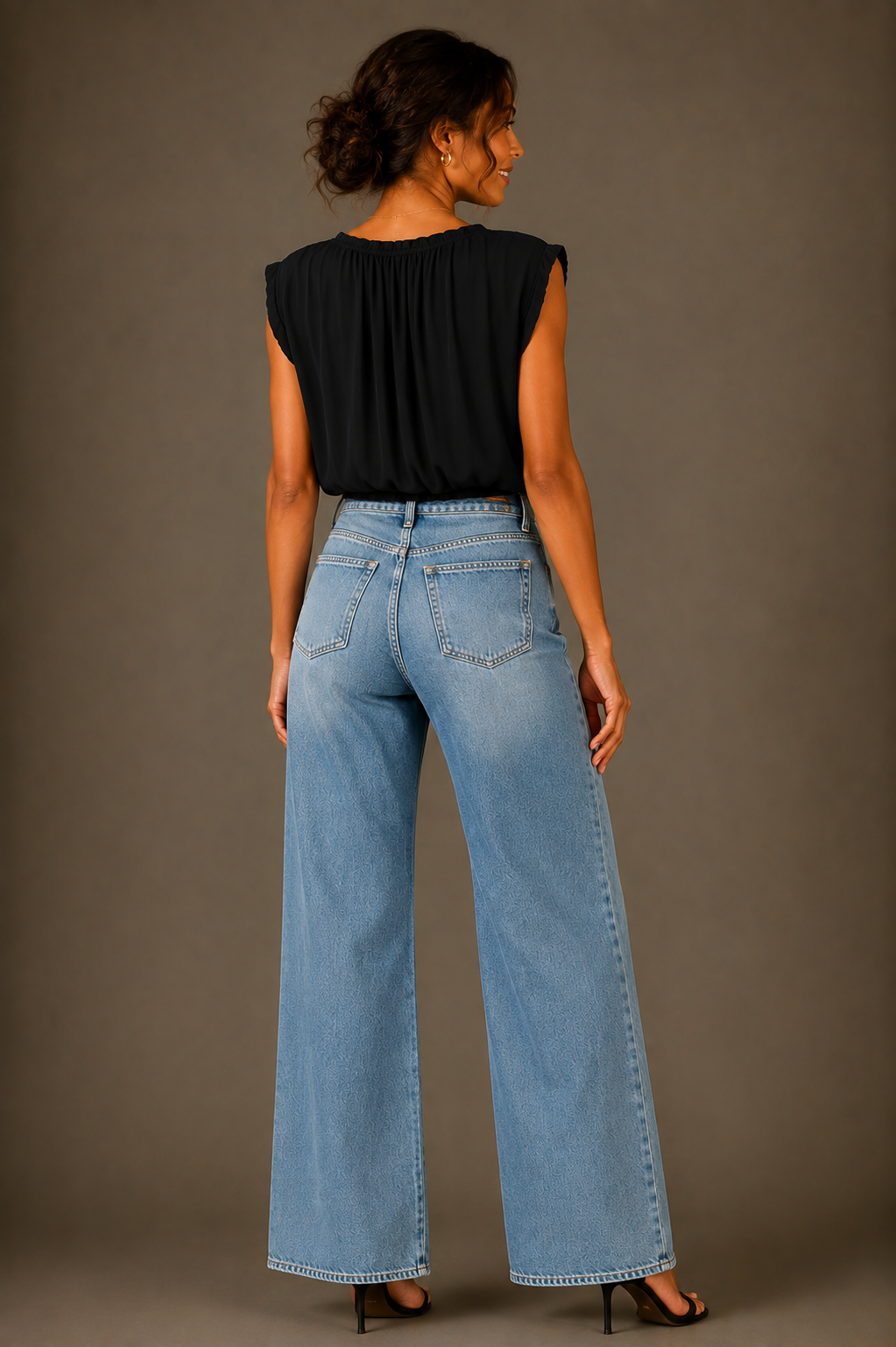 A woman shown from the back wearing a black sleeveless top and light blue wide-leg jeans, standing against a plain taupe studio background.