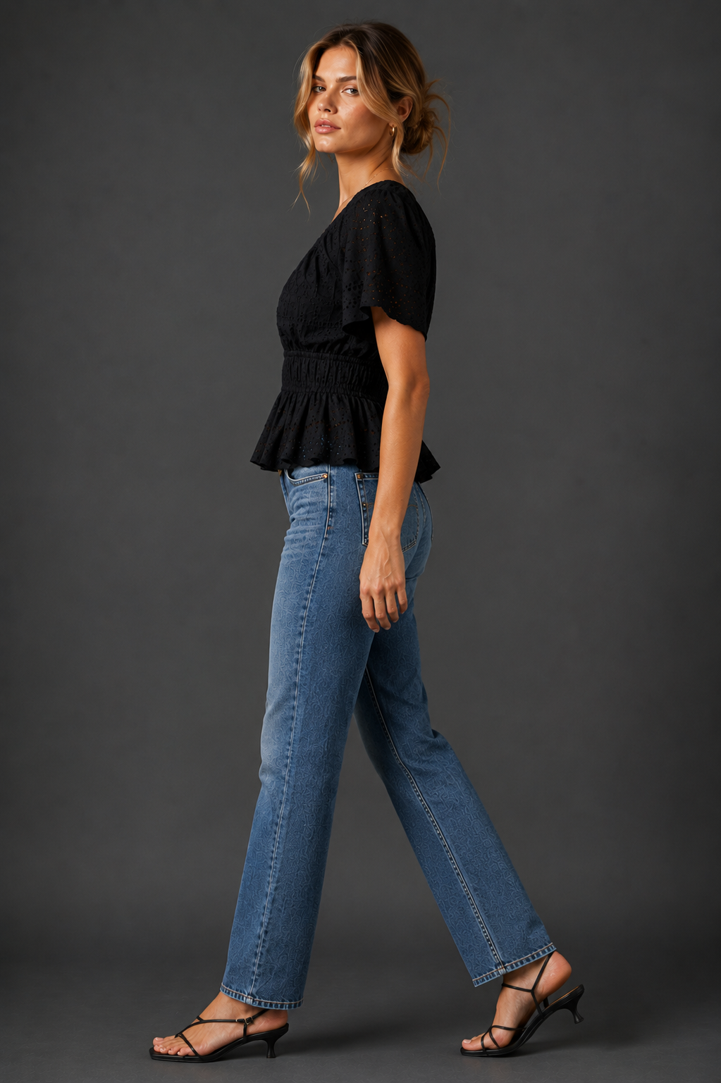 A woman modeling a black short-sleeve peplum top and blue straight-leg jeans against a dark gray studio background.