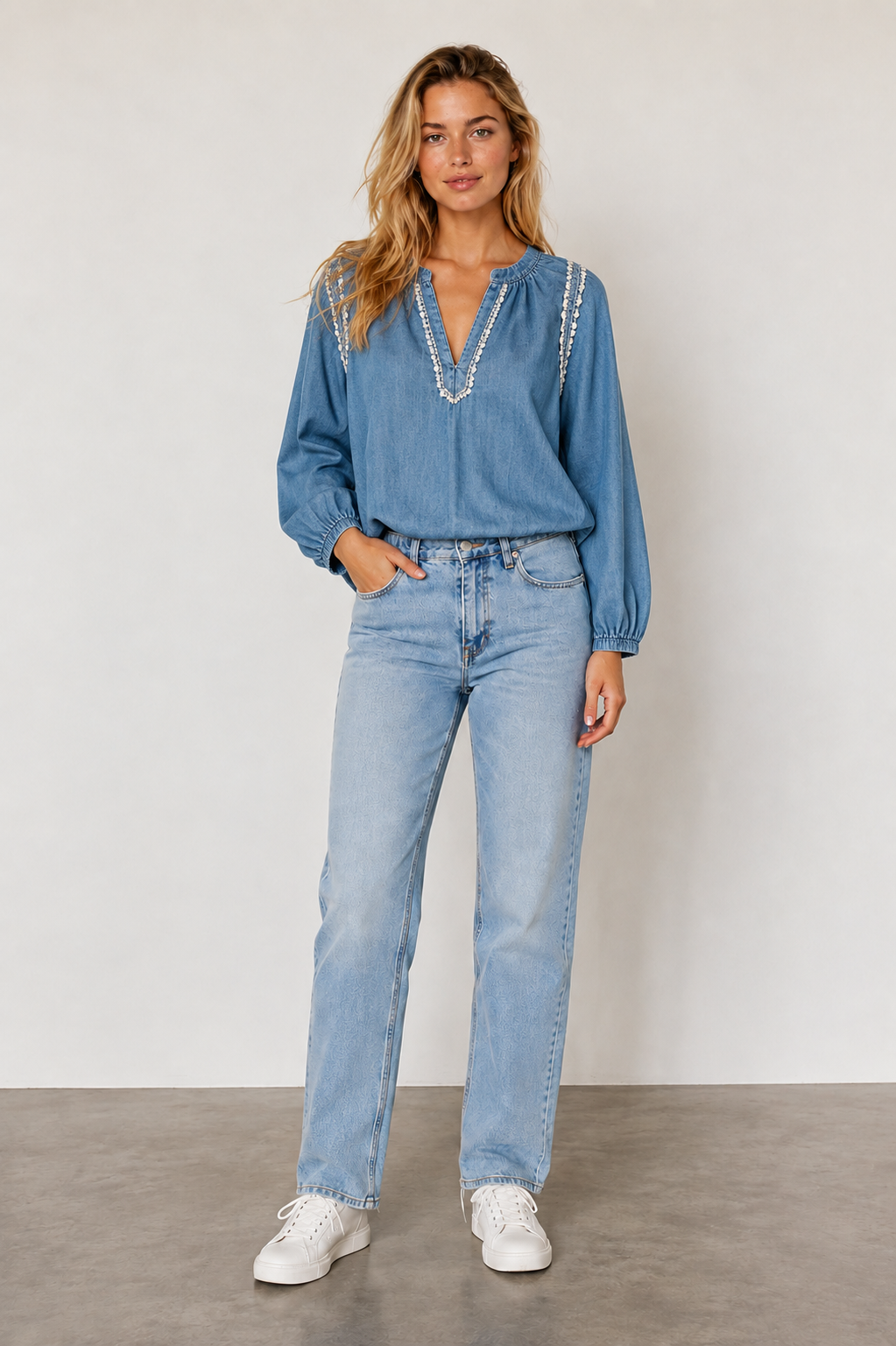 A woman modeling a light blue denim long-sleeve blouse with white beaded trim along the neckline, paired with light-wash blue jeans and white sneakers, standing against a neutral studio backdrop.
