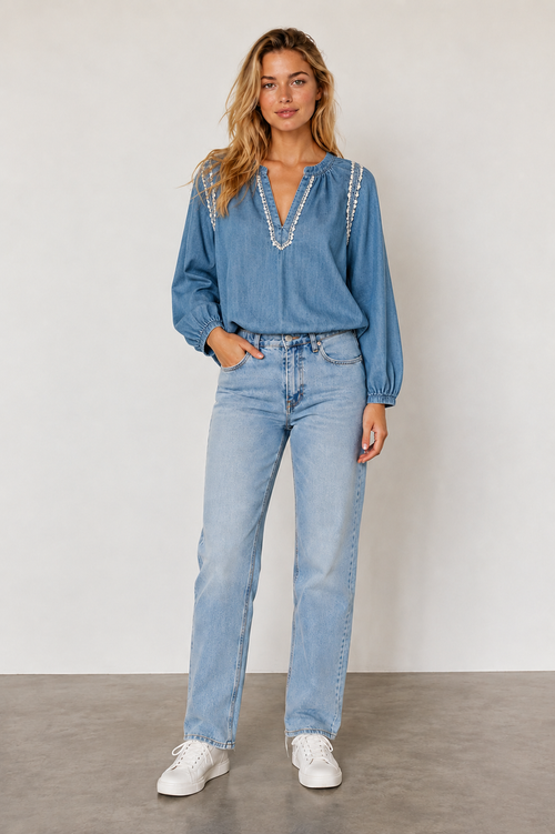 A woman modeling a light blue denim long-sleeve blouse with white beaded trim along the neckline, paired with light-wash blue jeans and white sneakers, standing against a neutral studio backdrop.