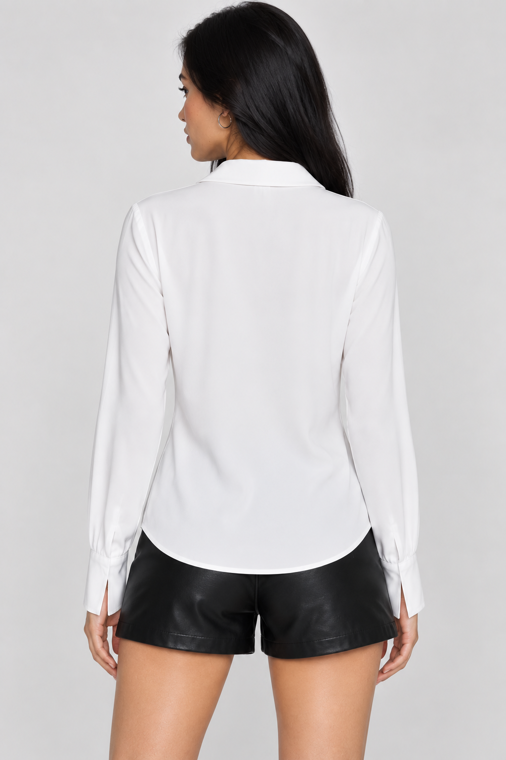 A woman shown from the back wearing a white long-sleeve button-up shirt and black faux-leather shorts against a light gray studio background.