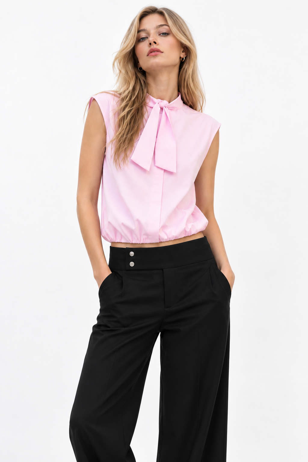 A woman modeling a sleeveless light pink blouse with a large bow tie-neck detail, paired with black wide-leg trousers, against a plain white studio background.