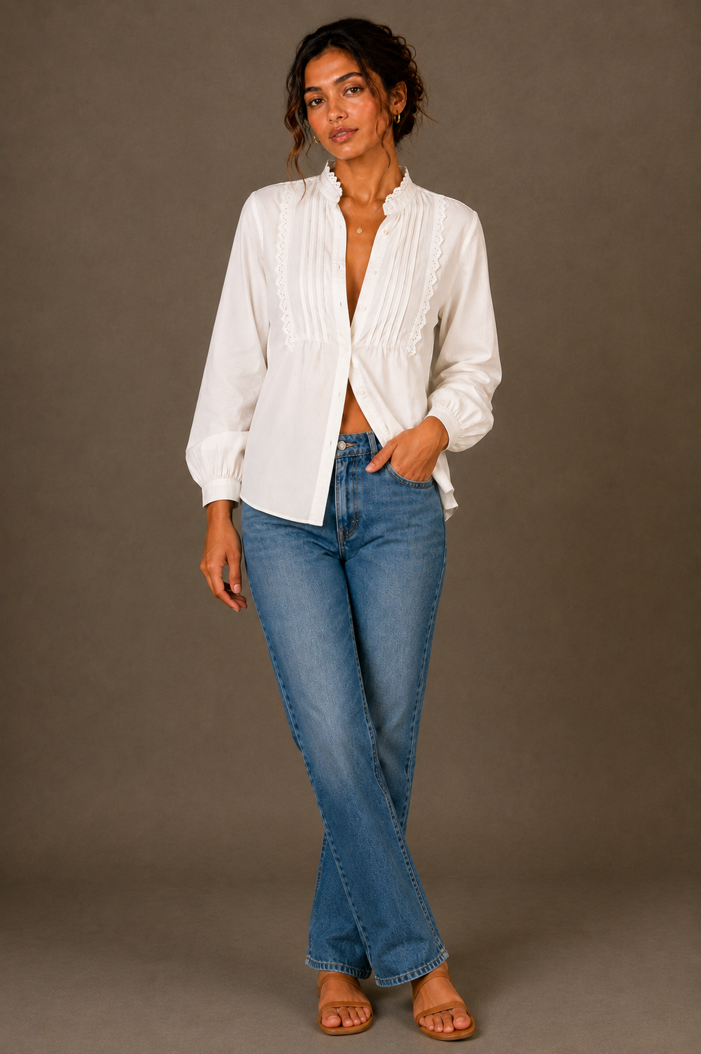 A woman modeling a white long-sleeve blouse with pleated front detailing and blue jeans, standing against a plain taupe studio background.