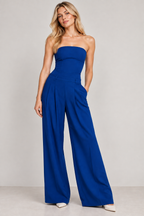 A woman modeling a strapless royal blue wide-leg jumpsuit against a plain light gray studio background.