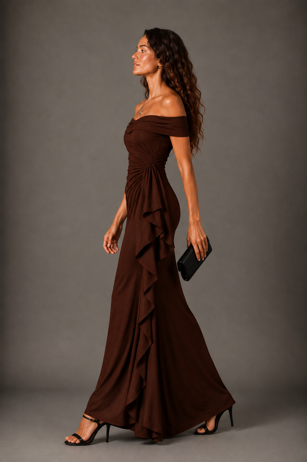 A woman modeling an off-the-shoulder, floor-length brown gown with ruffled layers along the side, paired with black high heels and holding a small black clutch, against a solid gray studio background.