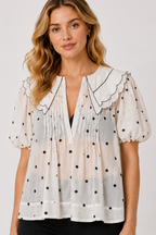 A woman wearing a sheer white short-sleeve blouse with black polka dots, a scalloped Peter Pan collar, and a smoky gradient hem, paired with black pants against a light gray studio background.