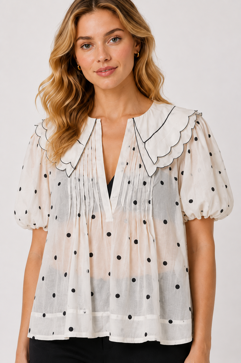 A woman wearing a sheer white short-sleeve blouse with black polka dots, a scalloped Peter Pan collar, and a smoky gradient hem, paired with black pants against a light gray studio background.