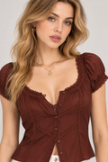 A woman wearing a brown puff-sleeve top with lace trim and a button-front bodice, photographed against a plain light gray studio background.