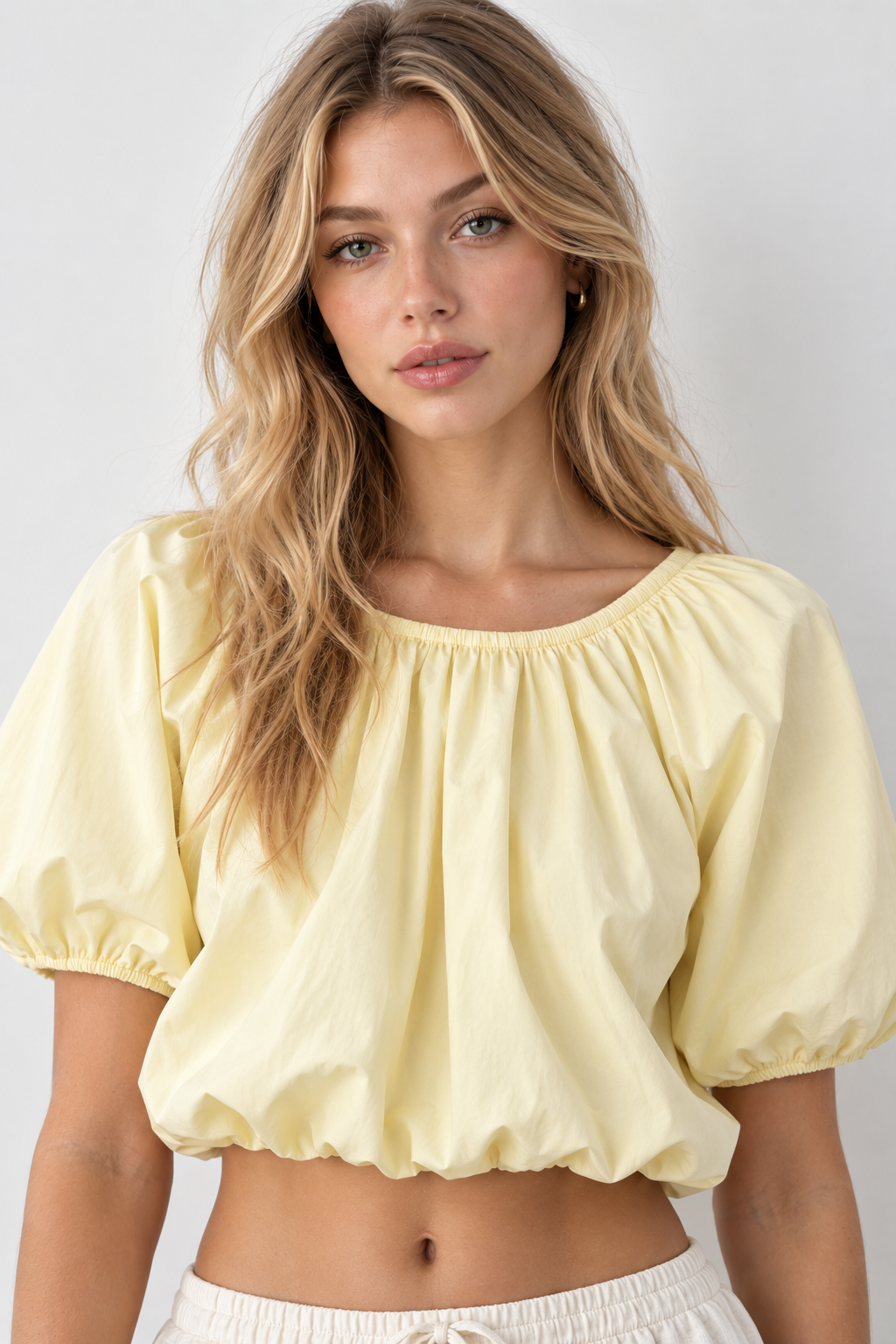 A woman wearing a pale yellow puff-sleeve crop top with a gathered neckline and elastic hem, paired with white drawstring shorts, against a light gray studio background.