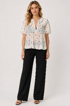 A woman modeling a white short-sleeve blouse with black polka dots and ruffled trim, paired with black wide-leg pants, against a light gray studio background.