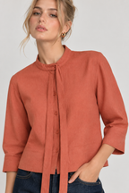 A woman wearing a terracotta-colored linen blouse with a tie-neck detail and three-quarter sleeves, paired with dark blue jeans, against a neutral light gray background.