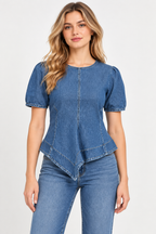 A woman modeling a blue denim short-sleeve top with puff sleeves and an asymmetrical ruffled hem, paired with blue jeans, against a plain light gray studio background.