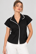 A woman modeling a black short-sleeve blouse with white pom-pom trim along the collar, placket, and sleeve edges, paired with white pants against a light gray studio background.