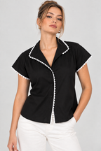 A woman modeling a black short-sleeve blouse with white pom-pom trim along the collar, placket, and sleeve edges, paired with white pants against a light gray studio background.